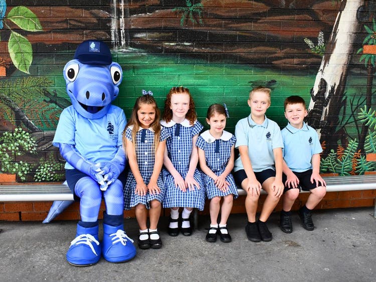 A group of 5 students sitting on a seat in front of a mural with Bluey, the school mascot.